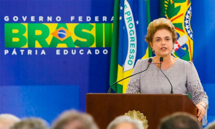 Dilma Rousseff discursando em primeiro plano. Ao fundo, a bandeira do Brasil e a logo do Governo Federal