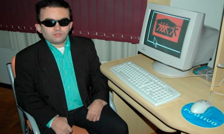 Homem sentado, de óculos escuros, ao lado de uma mesa com um computador branco