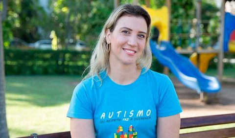 Mulher loira, sorrindo, sentada em um banco de praça, usando uma camisa azul com os dizeres: autismo: seu respeito faz a diferença