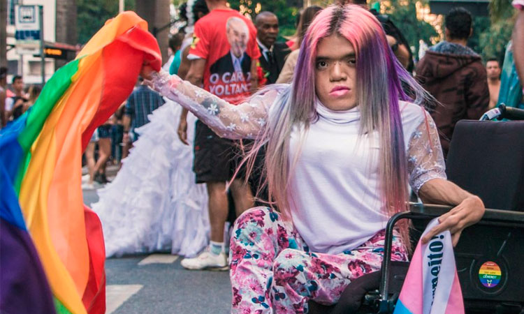 Travesti em uma cadeira de rodas, segurando uma bandeira do movimento LGBT, em meio a uma manifestação de rua