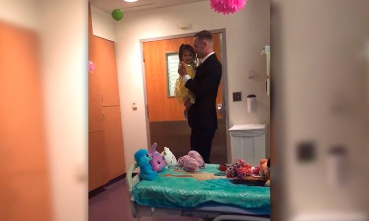 Pai e filha dançam em um quarto de hospital, com vários bichinhos de pelúcia em cima da cama. O pai veste um terno preto bastante elegante e a filha, em seu colo, usa um vestido amarelo de princesa.