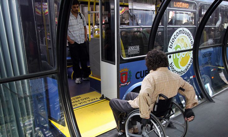 Homem em uma cadeira de rodas entra em um metrô da prefeitura de Curitiba, utilizando uma rampa amarela na porta. Na lateral do veículo, lê-se a placa redonda: 100% biocombustível.