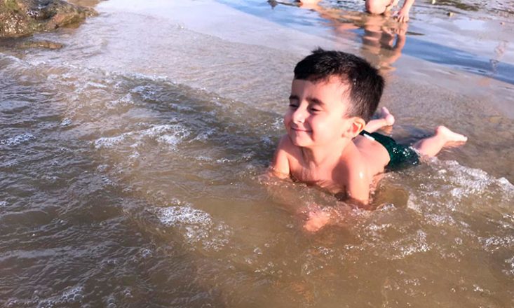 Foto horizontal de um garotinho de três anos brincando na água do mar, de bruços na areia