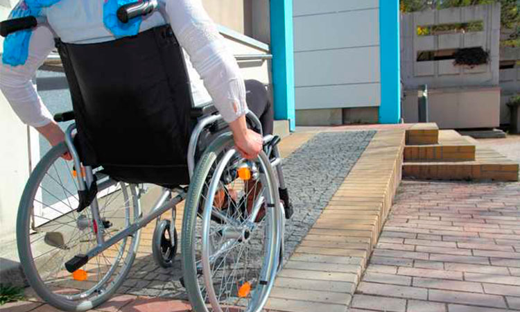 Homem em uma cadeira de rodas, do lado esquerdo da imagem, sobe a rampa de um prédio.