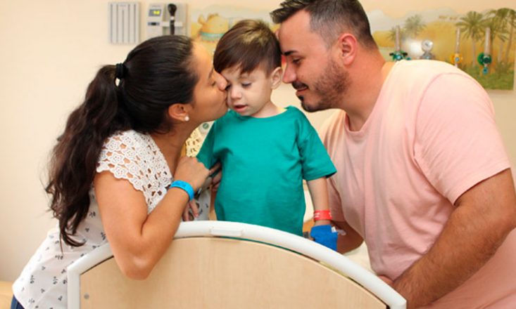 Foto horizontal de um garotinho em uma cama de hospital sendo beijado, de cada lado do rosto, por seus pais.
