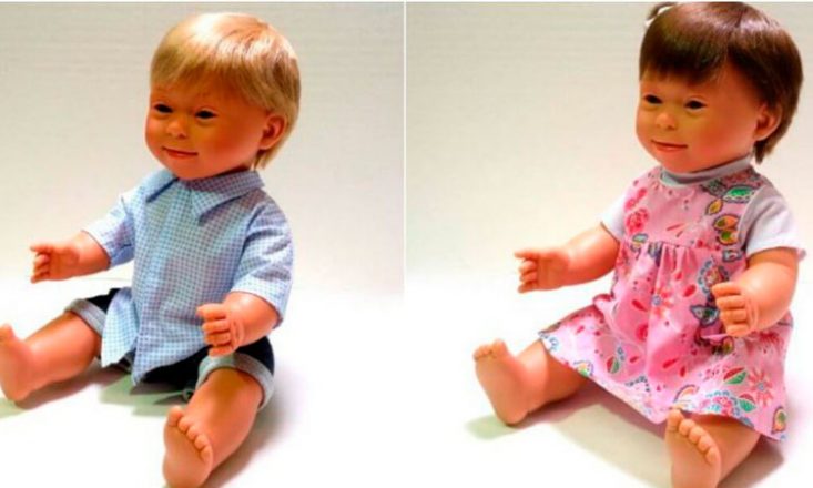 Duas bonecas lado a lado, ambas com aparência de Síndrome de Down. À esquerda, um menino loiro com uma camisa azul; à direita, uma menina morena em um vestido cor de rosa.