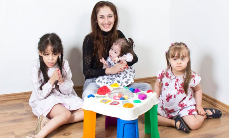 Uma mulher ao centro, com uma garotinha autista no colo, uma outra cega à esquerda e uma terceira, com Síndrome de Down, à direita. À frente, uma mesinha de brinquedo.
