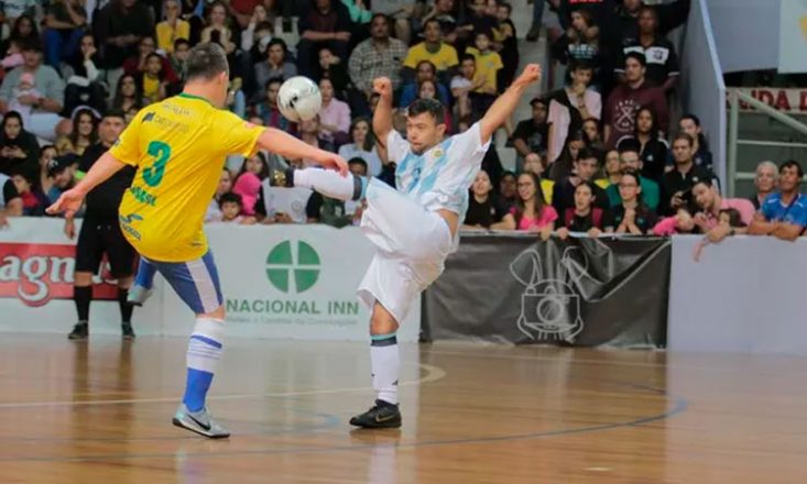 Dois rapazes com Síndrome de Down disputam uma jogada de futsal. À esquerda, o jogador veste a camisa amarela da seleção brasileira e, à direita, o jogador veste a camisa listrada em branco e azul da Argentina. Ao fundo, o juiz observa o lance e, atrás dele, arquibancada cheia de torcedores.