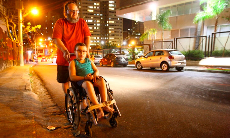 Pai e filho passeiam por uma rua de Belo Horizonte. O pai empurra a cadeira de rodas do garoto. Do lado direito da imagem, carros estacionados e luzes da rua, ao fundo.