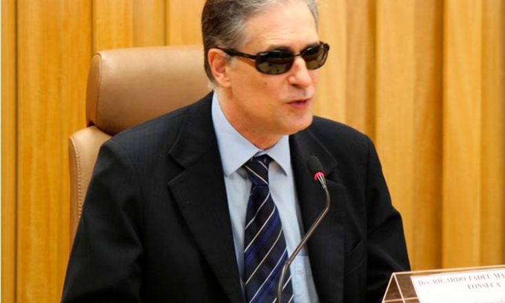 Homem cego, de óculos escuros e paletó preto, está sentado, discursando na frente de um microfone.