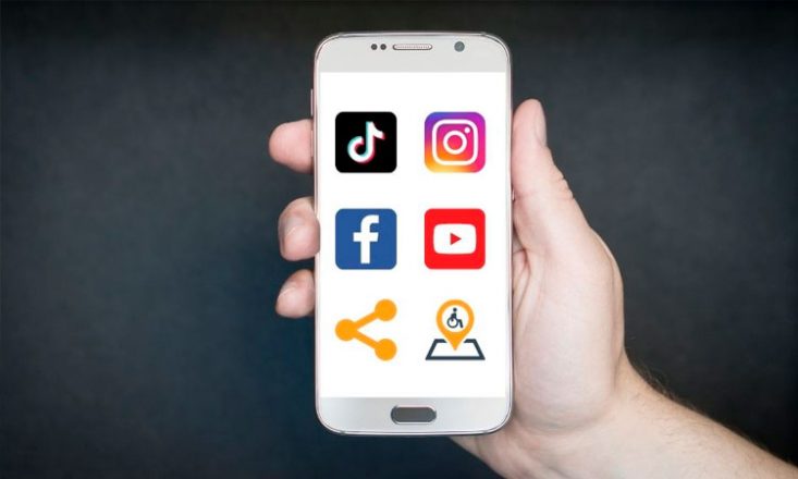 Foto horizontal da mão direita de um homem segurando um celular. Na tela, aparecem seis ícones - do Tiktok, Instagram, Facebook, Youtube, ícone de compartilhamento e do site Território Deficiente. Ao fundo, uma tela preta.